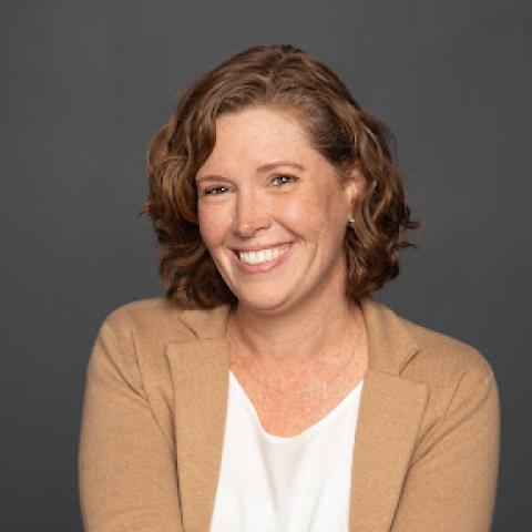 A woman with wavy brown hair wearing a tan blazer and white top smiles at the camera against a plain gray background.