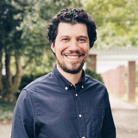 A man with curly dark hair and a beard, wearing a navy blue button-up shirt, smiles while standing outdoors with trees and a walkway in the background.