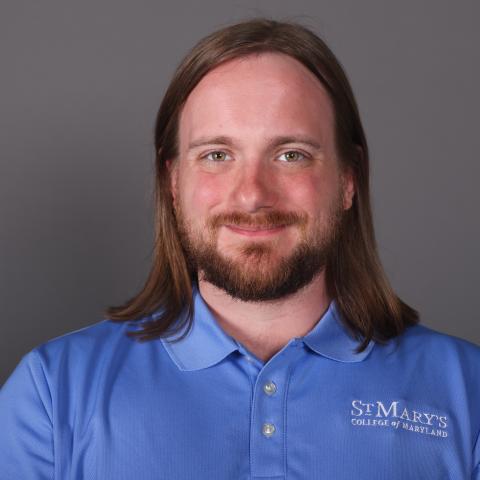 A person with long brown hair and a beard smiles at the camera, wearing a blue St. Mary’s College of Maryland polo shirt against a plain gray background.