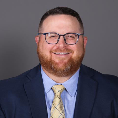 A man with short hair, a beard, and glasses, wearing a navy suit, light blue shirt, and yellow plaid tie, is smiling in front of a plain gray background.