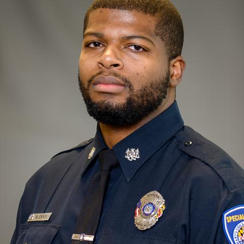A man in a police uniform with badges and patches poses for a formal portrait against a plain gray background.