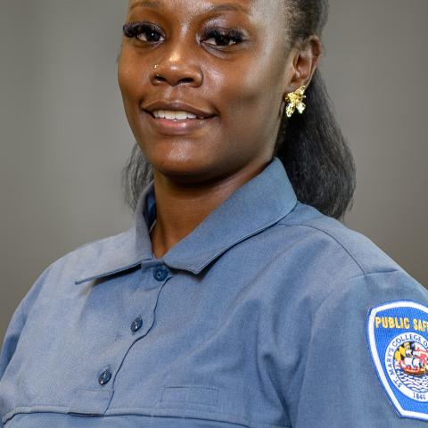 A woman in a blue public safety uniform with a badge on the shoulder, smiling, and standing against a plain background.