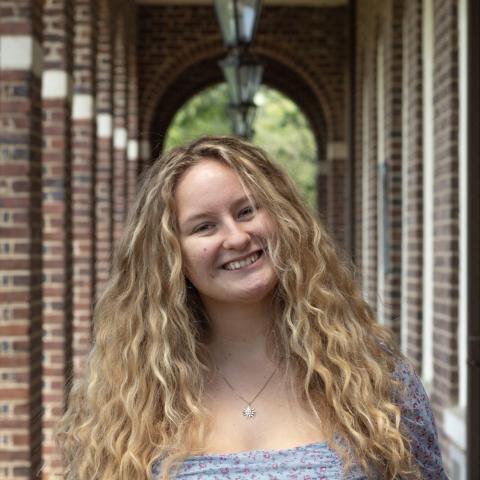 A young woman with long, curly blonde hair smiles at the camera while standing in a brick hallway with arches and hanging lanterns.