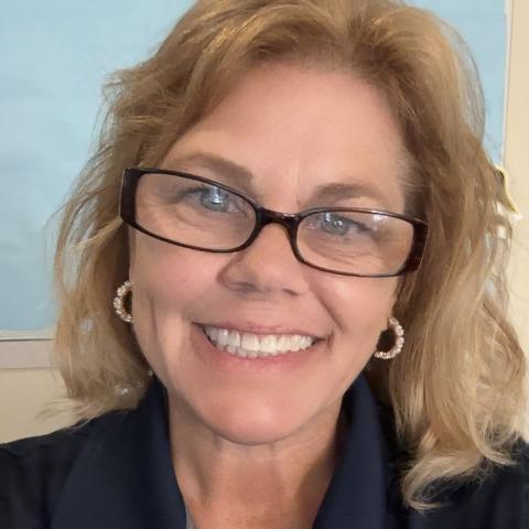 A woman with blonde hair and glasses smiles at the camera, wearing a navy collared shirt with a logo and the text "ST. MARY'S COLLEGE OF MARYLAND.
