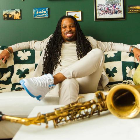 A man with long dreadlocks sits on a couch with arms outstretched, smiling, with a saxophone on the table in front of him and framed photos on the green wall behind.