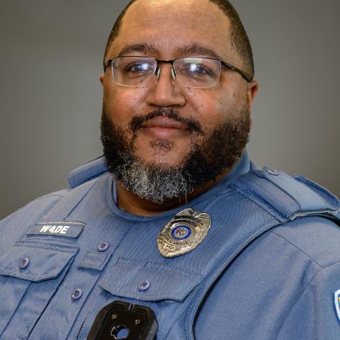 A police officer wearing glasses and a blue uniform with a badge and name tag that reads "WADE," poses for a portrait against a neutral background.