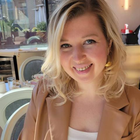 A woman with blonde hair, wearing a tan blazer and white top, smiles while sitting at a table in a sunlit café or restaurant.