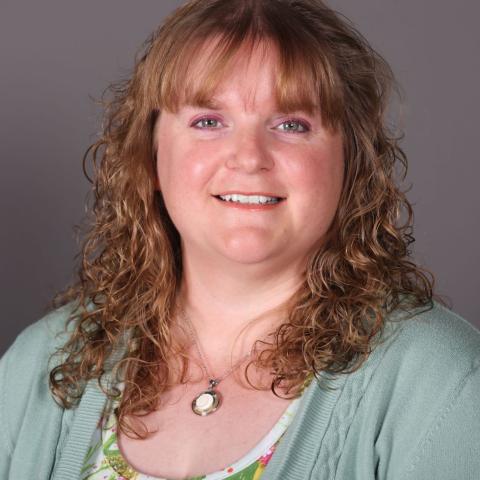 A woman with wavy light brown hair, wearing a green cardigan over a floral top, and a round pendant necklace, smiles at the camera against a plain background.