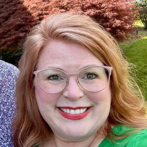 A woman with red hair, glasses, and a green top smiles outdoors with red and green foliage in the background.