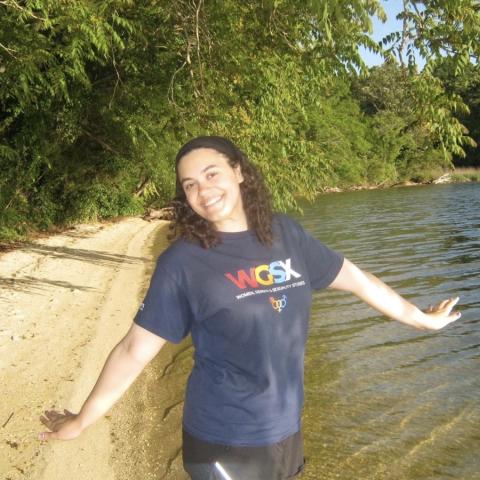 A person stands on a sandy lakeshore with arms outstretched, smiling, wearing a navy blue WGSX t-shirt, surrounded by green trees and water under a clear sky.