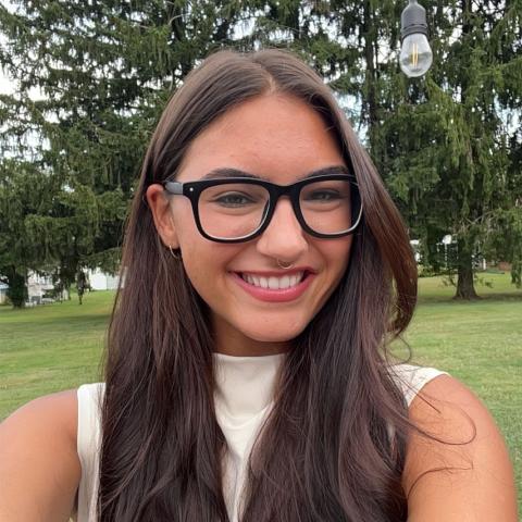 A woman with long brown hair and glasses smiles for a selfie outdoors, with green grass and tall trees in the background.