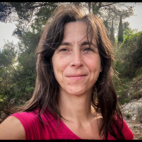 A woman with long brown hair wearing a pink shirt stands outdoors in a wooded area, smiling slightly at the camera. Trees and rocks are visible in the background.