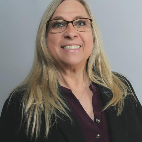A woman with long blonde hair and glasses, wearing a dark blazer over a purple blouse, smiles at the camera against a plain gray background.