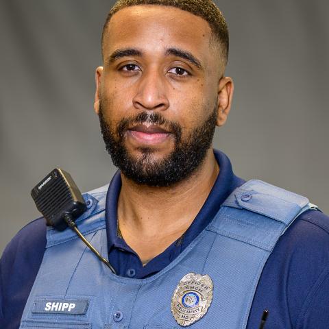 A police officer wearing a blue uniform and protective vest with a name tag reading "Shipp" stands facing the camera, a radio microphone clipped to his shoulder.