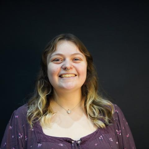A person with long, wavy hair and a nose ring is smiling while standing in front of a plain dark background.