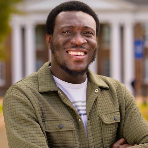 A man with short hair and a beard, wearing a green quilted jacket and smart watch, smiles with arms crossed in front of a building with white columns.