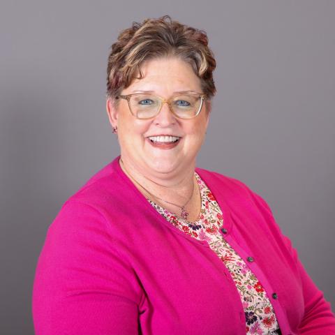A woman with short, wavy hair wearing glasses, a bright pink cardigan, and a floral blouse, smiling in front of a plain gray background.