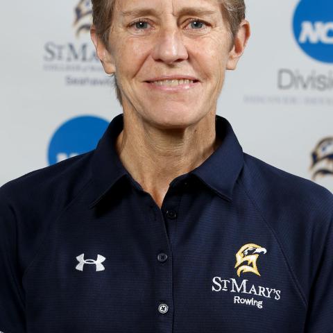 A person with short gray hair wearing a navy blue St. Mary's Rowing polo shirt stands in front of a backdrop with NCAA and St. Mary's logos.