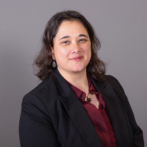 A person with shoulder-length dark hair wearing a black blazer, maroon blouse, and necklace poses for a professional portrait against a gray background.