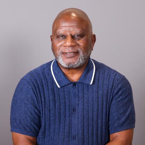 A middle-aged man with a gray beard wearing a blue short-sleeve polo shirt sits in front of a plain gray background, smiling slightly at the camera.