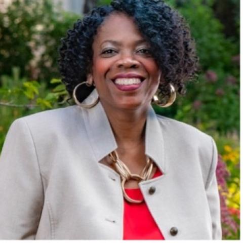 Woman with short curly hair, wearing a light beige blazer, red top, and large gold hoop earrings, smiling outdoors with greenery in the background.