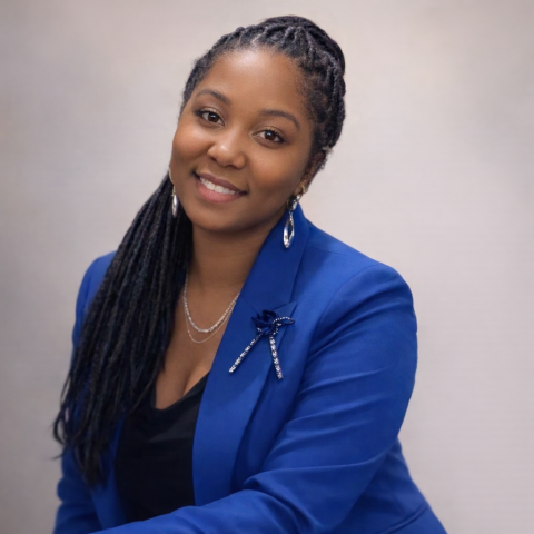 A woman with long braided hair wears a bright blue blazer, a black top, and silver jewelry, smiling at the camera.