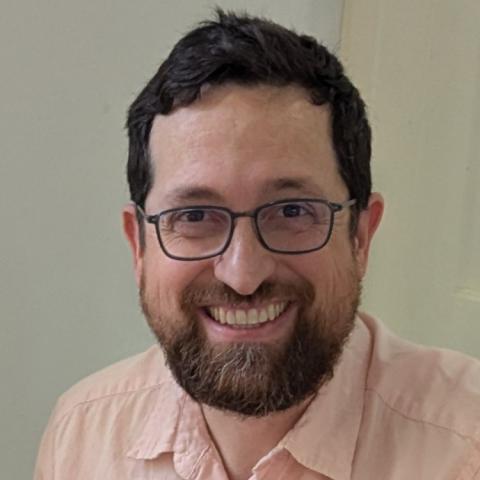 A man with dark hair, glasses, and a beard smiles at the camera while wearing a light pink collared shirt.