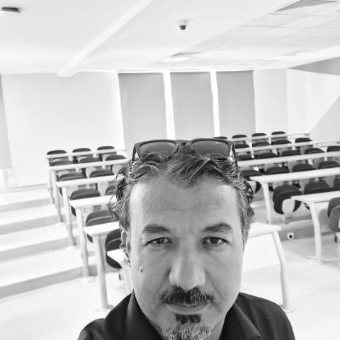 A man stands in the foreground of an empty classroom with rows of desks and chairs; the photo is in black and white.