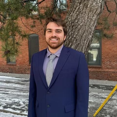 A man in a blue suit and gray tie stands outside on a snowy sidewalk in front of a brick building and a large tree.