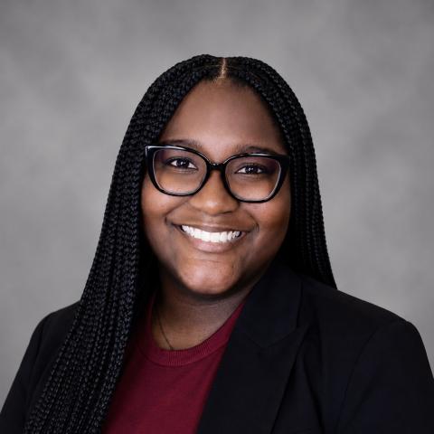 A woman with long braided hair, glasses, and a black blazer over a maroon shirt smiles at the camera against a gray background.