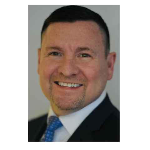 A man with short dark hair, a trimmed goatee, and a suit with a blue polka dot tie smiles at the camera against a neutral background.