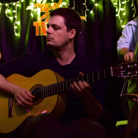 A man plays an acoustic guitar on stage with other musicians; string lights hang above them in a dimly lit venue.