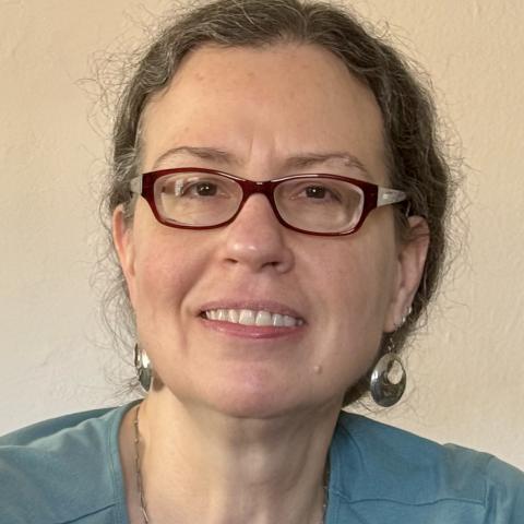 A woman with brown hair with some grey, wearing glasses, earrings and a teal top, smiling at the camera in front of a light-colored wall.