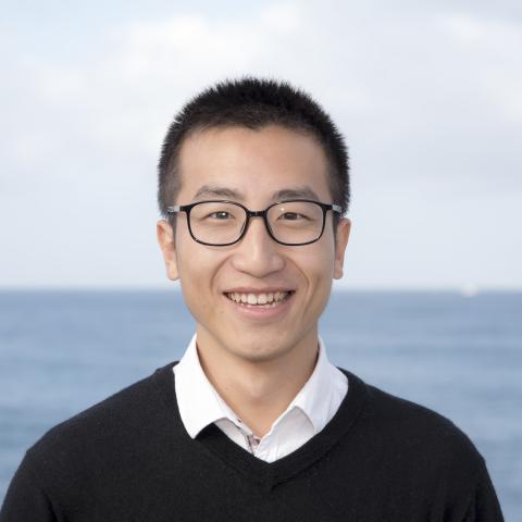 A person wearing glasses, a white collared shirt, and a black sweater smiles while standing in front of a body of water with a cloudy sky in the background.