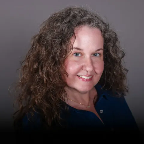 Woman with curly brown hair, blue eyes, and a blue shirt smiles at the camera against a plain gray background.