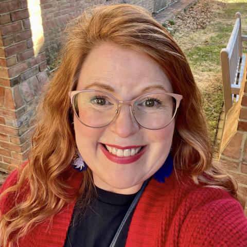 A woman with long red hair, wearing glasses, a red sweater, and blue earrings, smiles outdoors near a brick building and lawn.