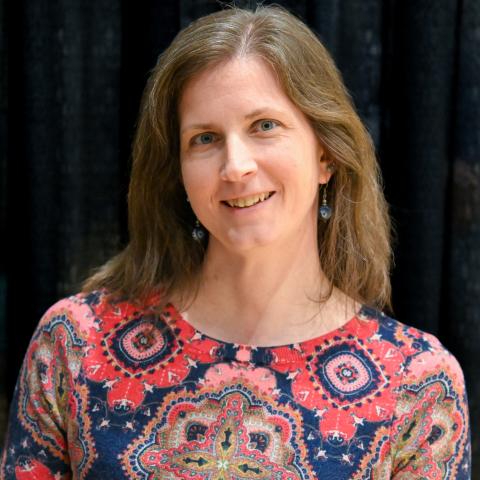 A woman with shoulder-length brown hair, wearing a patterned red and blue top, stands in front of a dark curtain, smiling at the camera.