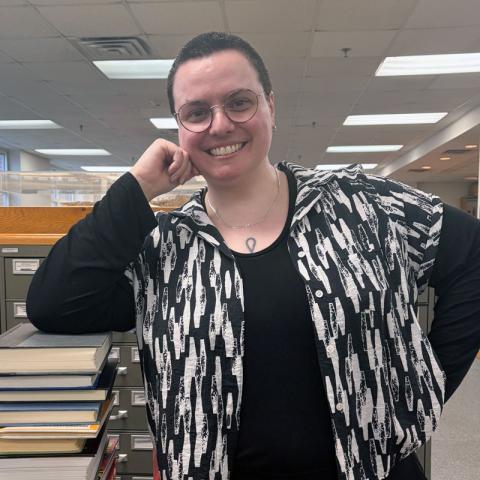 A person with short hair and glasses smiles while leaning on a file cabinet in a library, wearing a black and white patterned shirt over a black top.