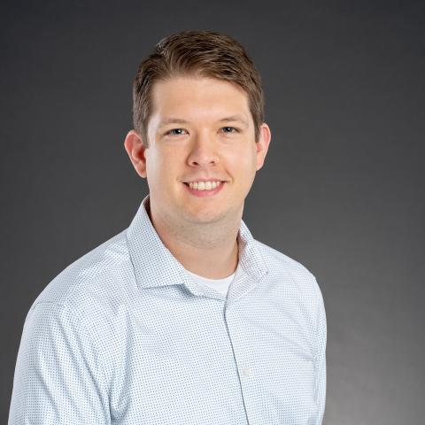 Man with short brown hair wearing a light blue button-up shirt poses in front of a plain dark gray background, smiling at the camera.