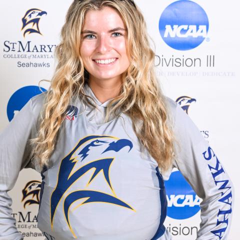 Young woman smiling, wearing a Seahawks athletic hoodie and sunglasses on her head, standing in front of a St. Mary’s College of Maryland and NCAA Division III step-and-repeat banner.