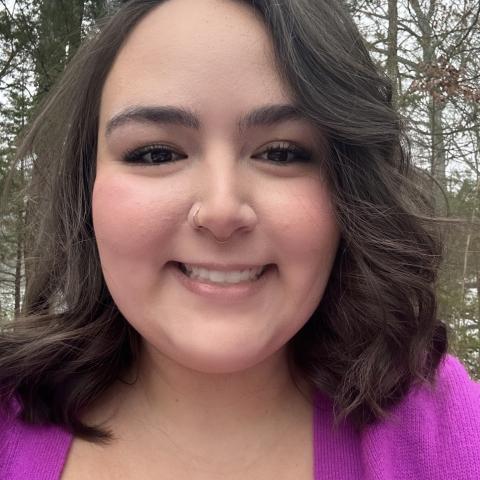 A person with shoulder-length dark hair, wearing a purple top and a nose ring, smiles outdoors with trees in the background.