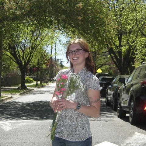 A person with glasses stands in the street holding pink flowers, surrounded by cars and leafy trees on a sunny day.