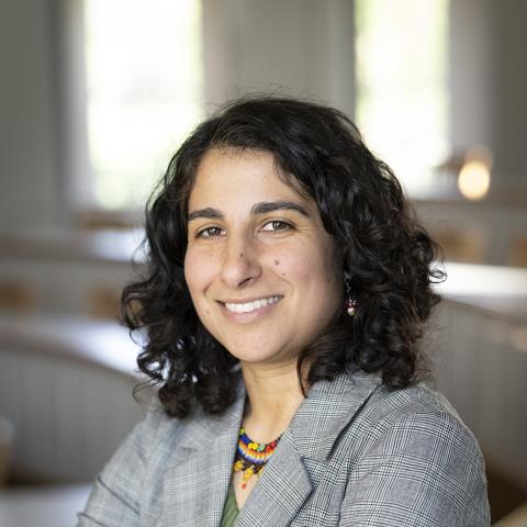 A person with curly dark hair wearing a plaid blazer and colorful necklace stands in a bright, empty classroom.