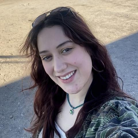 A woman with long brown hair in a camouflage jacket and sunglasses on her head smiles outdoors on a sunny day.
