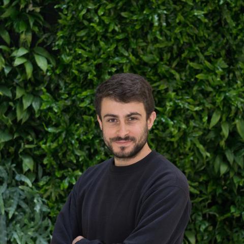 A man with short dark hair and a beard, wearing a black sweatshirt, stands in front of a lush green leafy background, looking at the camera with a neutral expression.