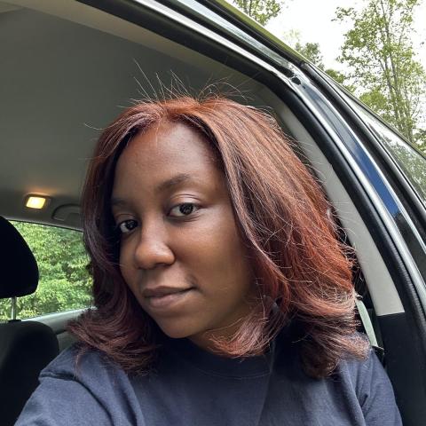 Woman with shoulder-length reddish-brown hair sits in a car with the door open, wearing a dark shirt. Trees and greenery are visible outside.