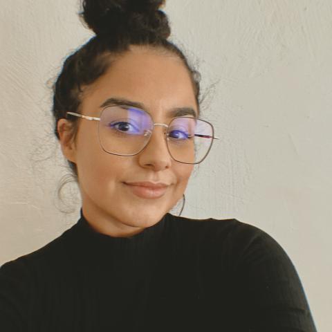 A woman with glasses and a black top in front of a plain light-colored wall.