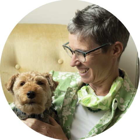 A person with short gray hair and glasses smiles while holding a small, brown dog on a beige chair.
