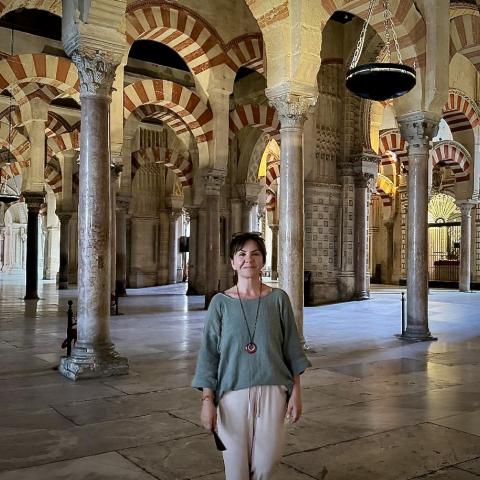 A person stands inside a historic building with striped arches, marble columns, and patterned walls, illuminated by natural light.