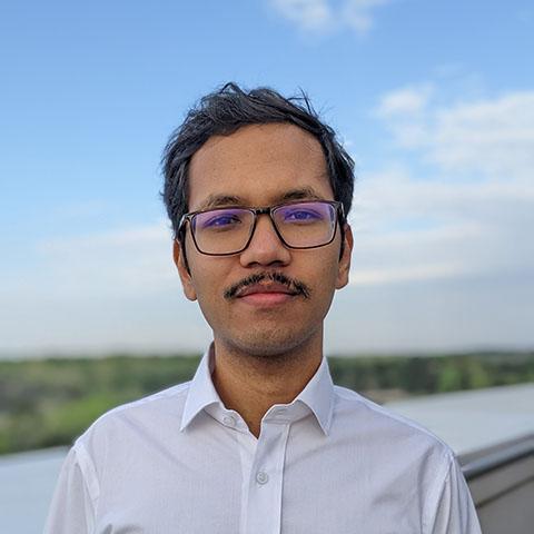 A man with glasses and a mustache, wearing a white shirt, stands outdoors with a blurred background of greenery and a partly cloudy sky.